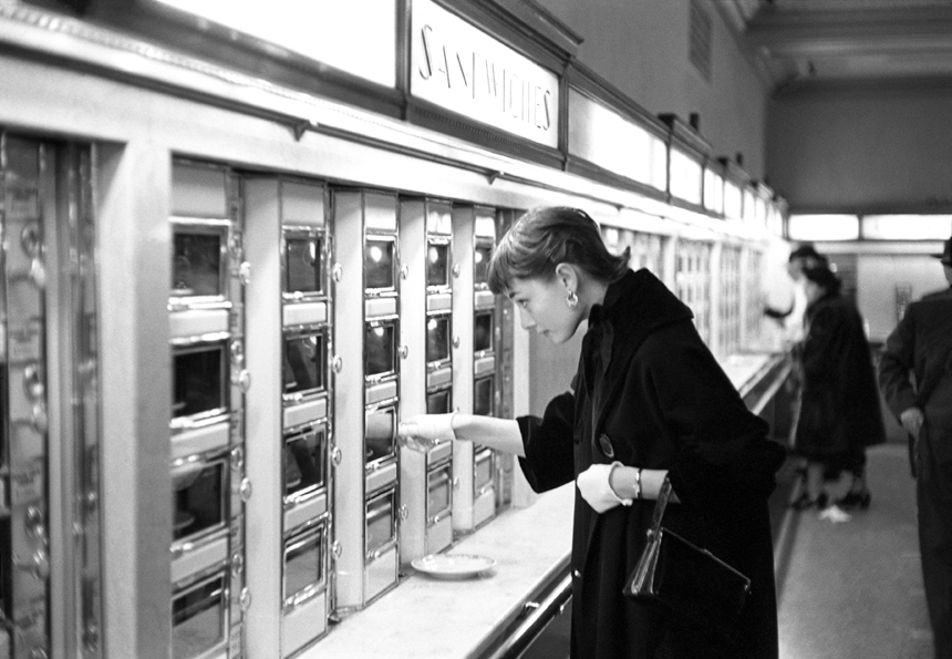 Audrey Hepburn is at an automat and opening a compartment to grab a sandwich.